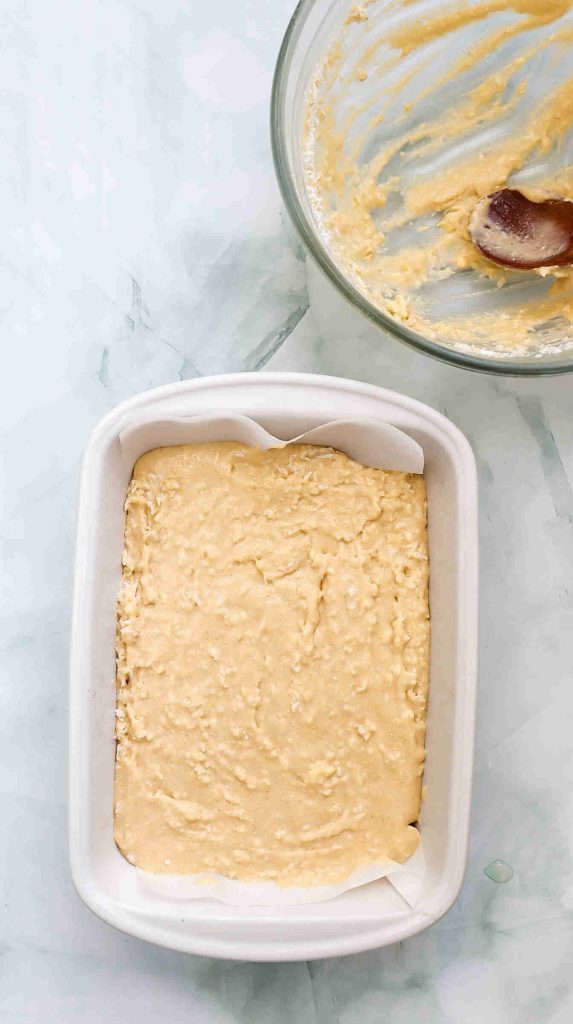 A rectangular white baking dish filled with unbaked coconut Christmas cake batter sits on a marble surface. Nearby is a glass bowl with leftover batter and a wooden spoon.