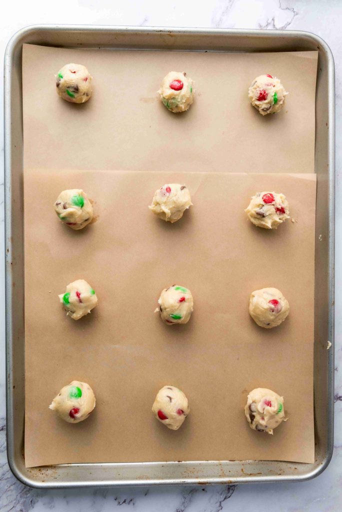 A baking sheet lined with parchment paper holds twelve evenly spaced scoops of Christmas M&M cookies dough, each packed with festive red and green candies, ready to be baked.