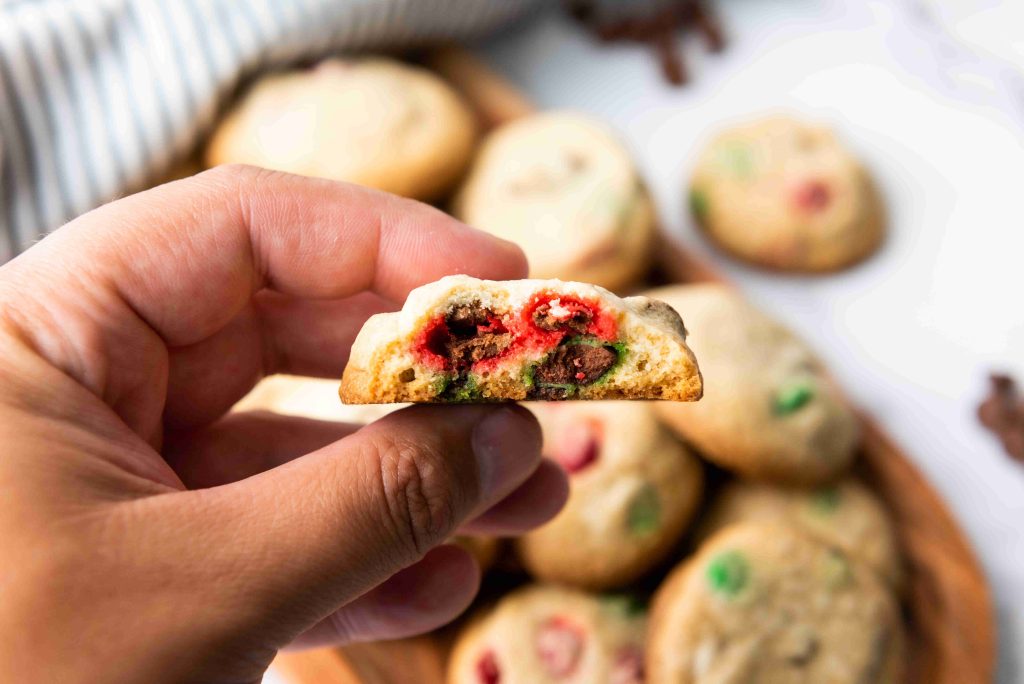 A hand holds a half-eaten Christmas M&M cookie, revealing a soft inside with festive red and green candies. Several whole cookies with colorful M&Ms are blurred in the background.