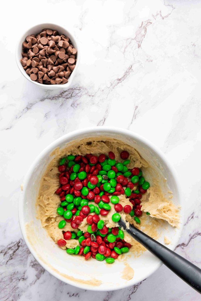 A white bowl of cookie dough mixed with red and green Christmas M&M cookies sits on a marble surface. A black spoon rests in the bowl, while a nearby small bowl holds chocolate chips.