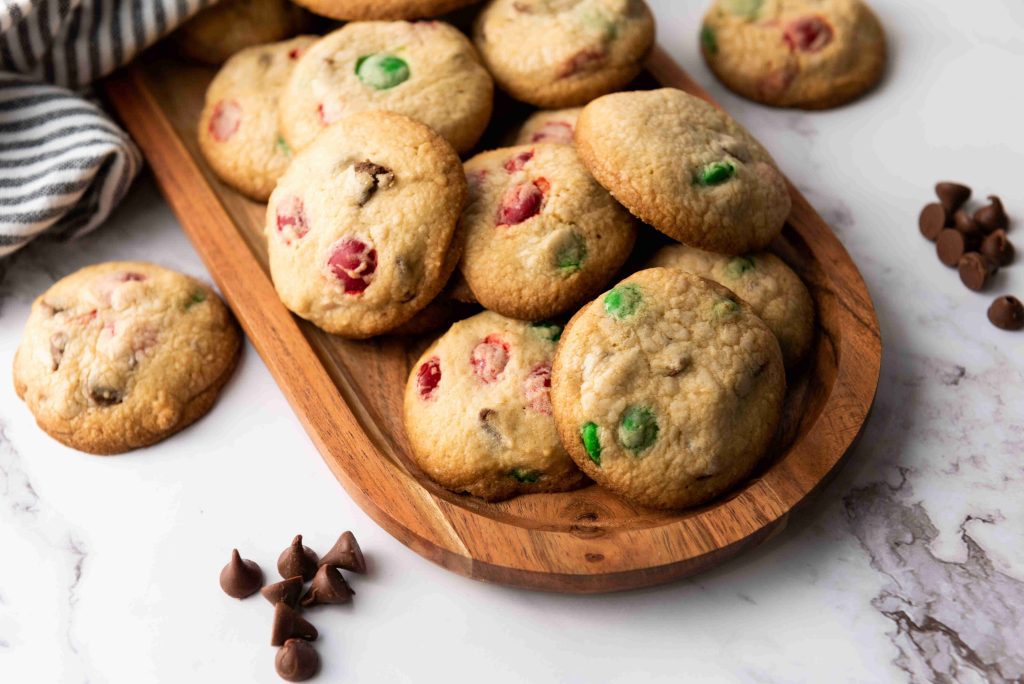 A wooden tray filled with Christmas M&M cookies, studded with festive red and green candy pieces, sits on a marble surface, with a few cookies and chocolate chips scattered nearby.