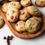 A wooden tray filled with Christmas M&M cookies, studded with festive red and green candy pieces, sits on a marble surface, with a few cookies and chocolate chips scattered nearby.