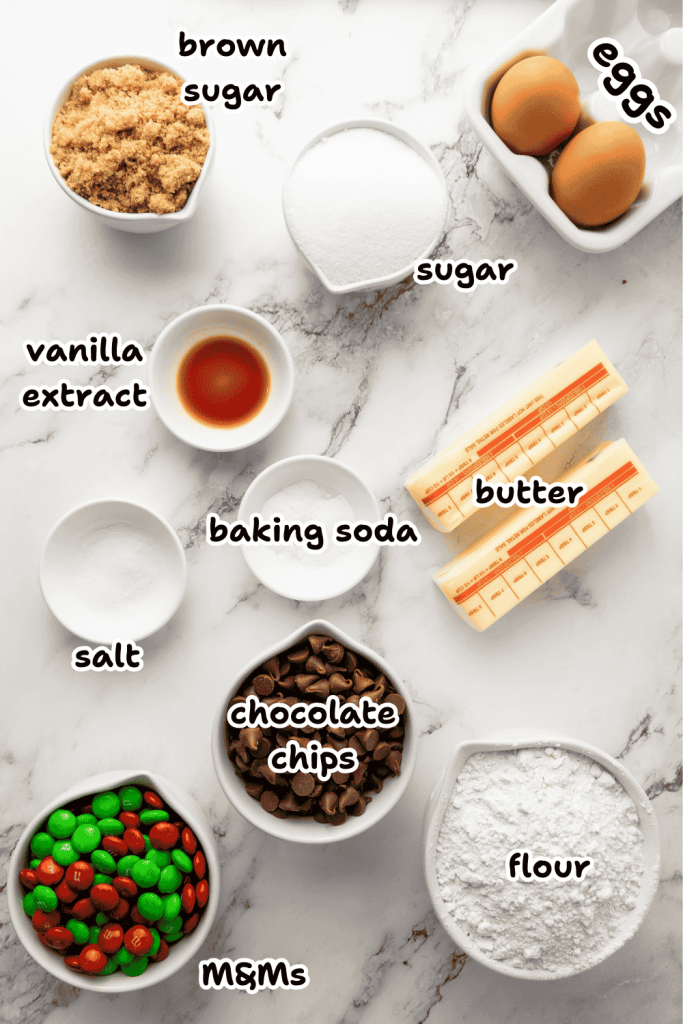 A marble countertop displays labeled baking ingredients in bowls for Christmas M&M cookies: brown sugar, white sugar, eggs, vanilla extract, baking soda, salt, butter sticks, chocolate chips, flour, and festive M&Ms.