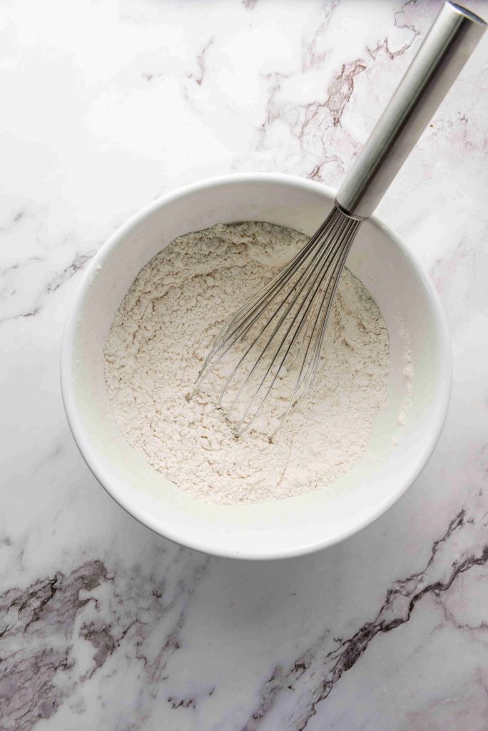 A metal whisk rests in a white bowl filled with flour, ready to bake Christmas MM cookies, on a white and gray marble countertop.