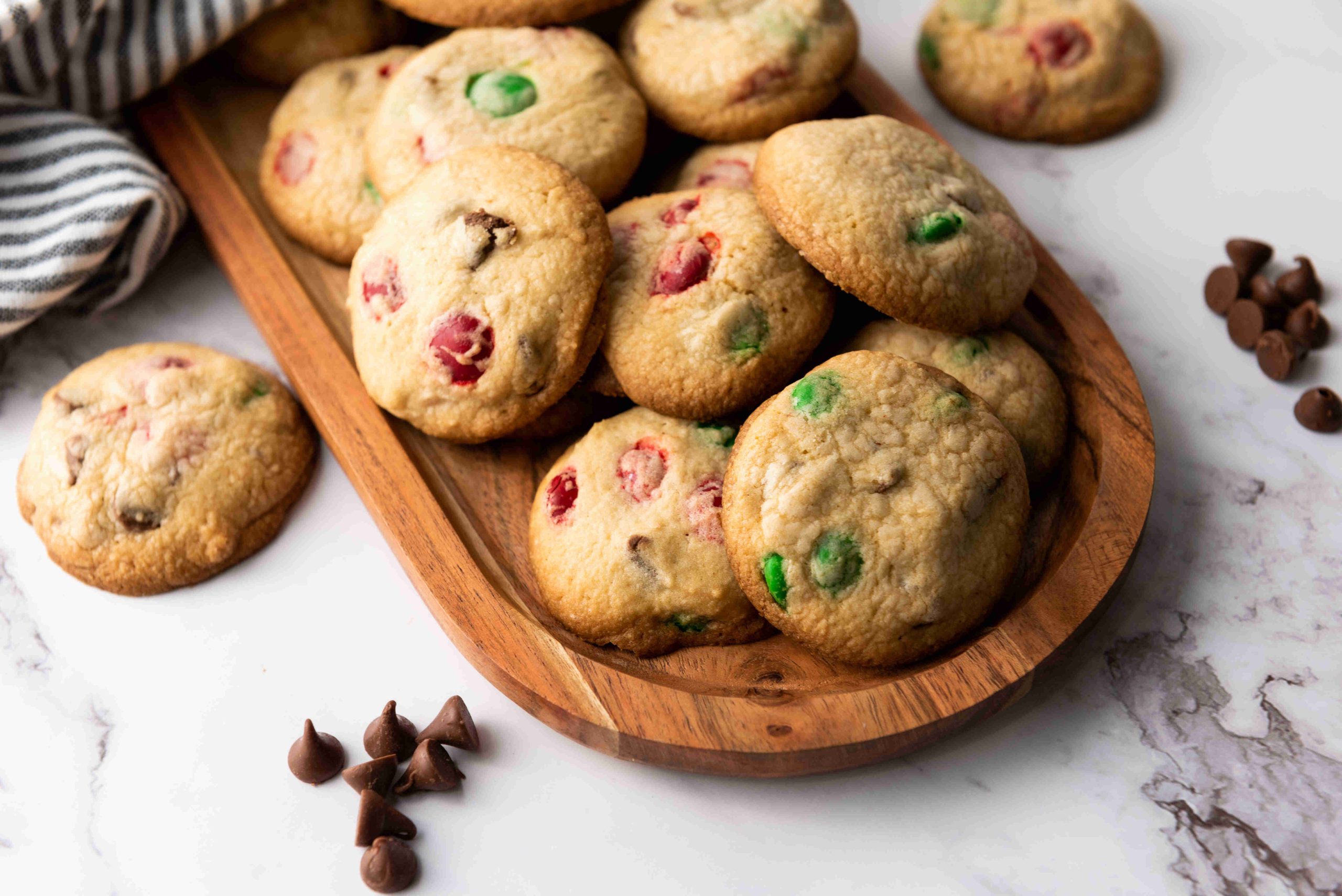 A wooden tray filled with Christmas M&M cookies, studded with festive red and green candy pieces, sits on a marble surface, with a few cookies and chocolate chips scattered nearby.