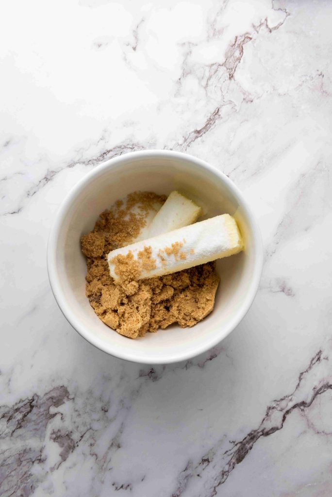 A white bowl containing two sticks of butter and brown sugar, perfect for making Christmas MM cookies, sits on a white marble surface.