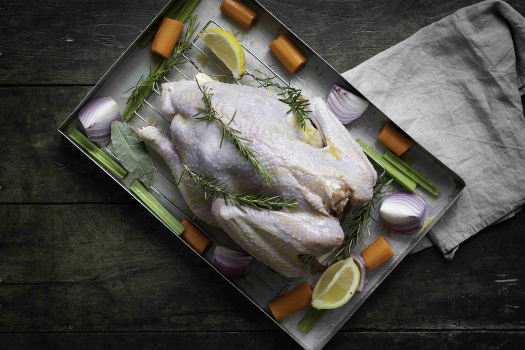 Raw whole chicken in a roasting pan with rosemary, lemon wedges, and surrounded by chopped carrots, celery, onions, and bay leaves—like a holiday turkey—ready for cooking on a dark wooden surface with a gray cloth beside the pan.