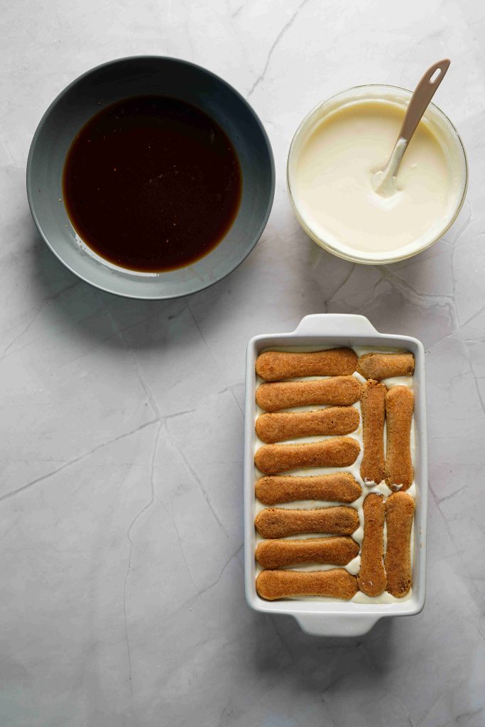 Overhead view of a dish with ladyfinger biscuits and cream, a bowl of coffee, and a bowl of cream with a spatula on white marble, showing Christmas tiramisu ingredients and assembly.