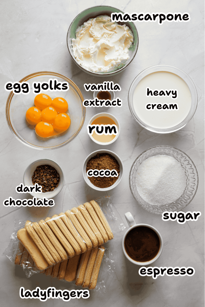 Ingredients for Christmas tiramisu are arranged on a white surface: mascarpone, egg yolks, vanilla extract, rum, heavy cream, cocoa, dark chocolate, sugar, espresso, and ladyfingers. Each festive item is clearly labeled.