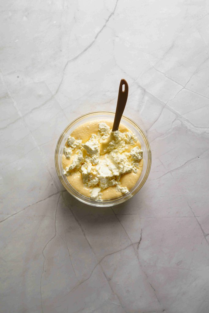 A glass bowl filled with creamy yellow Christmas tiramisu, topped with dollops of white whipped cream, sits on a light marble surface. A small brown spoon rests in the bowl.