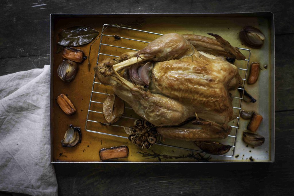 A whole roasted chicken sits on a rack in a roasting pan, surrounded by holiday turkey-style roasted vegetables like carrots, onions, and garlic. A white cloth rests beside the pan on a dark wooden surface.