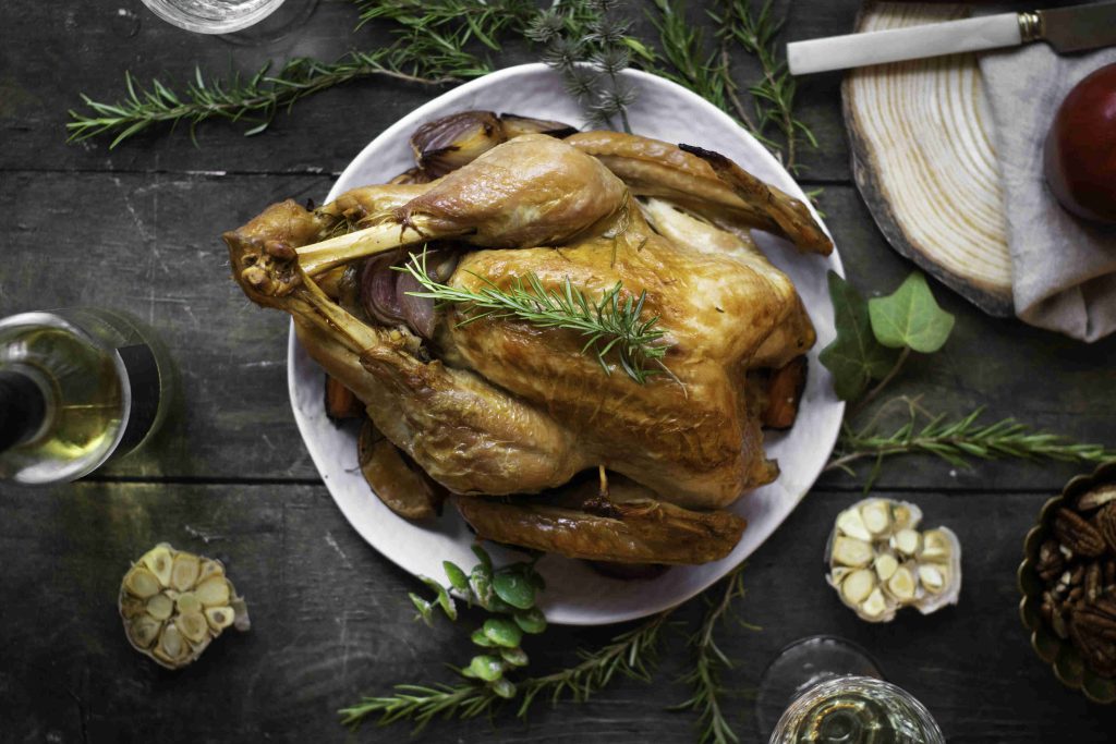 A roasted whole chicken garnished with fresh rosemary sits on a white plate. The table, reminiscent of a Holiday Turkey feast, is decorated with herbs, garlic halves, wine, and rustic wooden elements for a festive atmosphere.