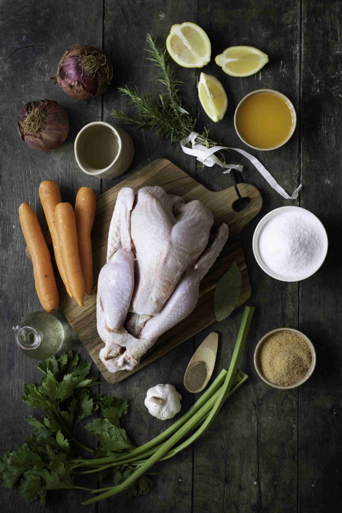 A whole raw chicken on a wooden board surrounded by carrots, onions, lemon wedges, fresh herbs, garlic, green onions, sugar, brown sugar, and a cup of broth—perfect for those who prefer chicken over Holiday Turkey.