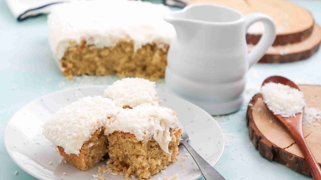 A plate with three square pieces of coconut Christmas cake topped with white frosting and shredded coconut, with a larger cake, a pitcher, and a wooden spoon of coconut flakes in the background.