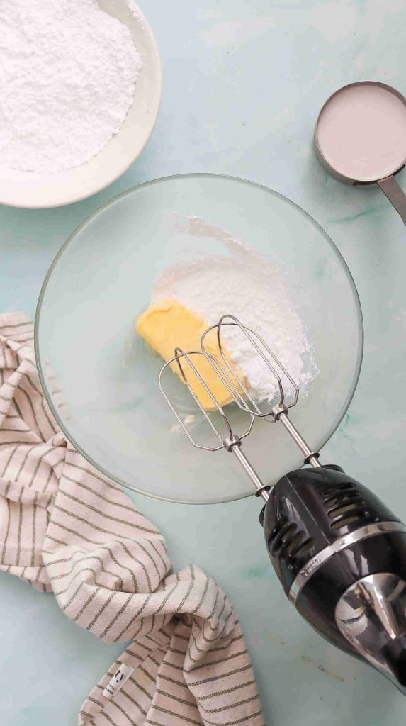 A stand mixer with beaters is positioned in a glass bowl containing a stick of butter and powdered sugar—perfect for starting a coconut Christmas cake. Nearby are a striped kitchen towel, a plate of powdered sugar, and a metal measuring cup on a light blue surface.