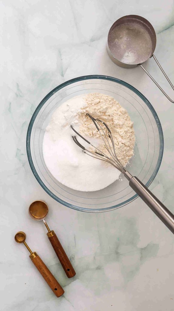 A glass bowl with flour and sugar for coconut Christmas cake is being mixed with a metal whisk. Nearby are a metal measuring cup and two wooden-handled measuring spoons on a light marble surface.
