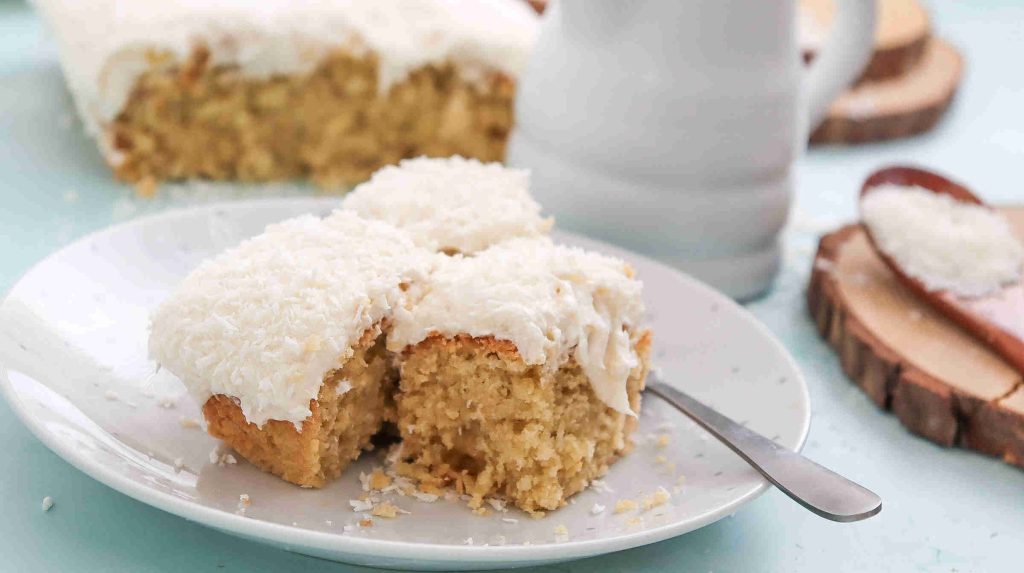 Three pieces of coconut Christmas cake with white frosting and shredded coconut on top are served on a white plate, with a fork beside them. More cake and a jar are in the background.