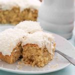 Three pieces of coconut Christmas cake with white frosting and shredded coconut on top are served on a white plate, with a fork beside them. More cake and a jar are in the background.