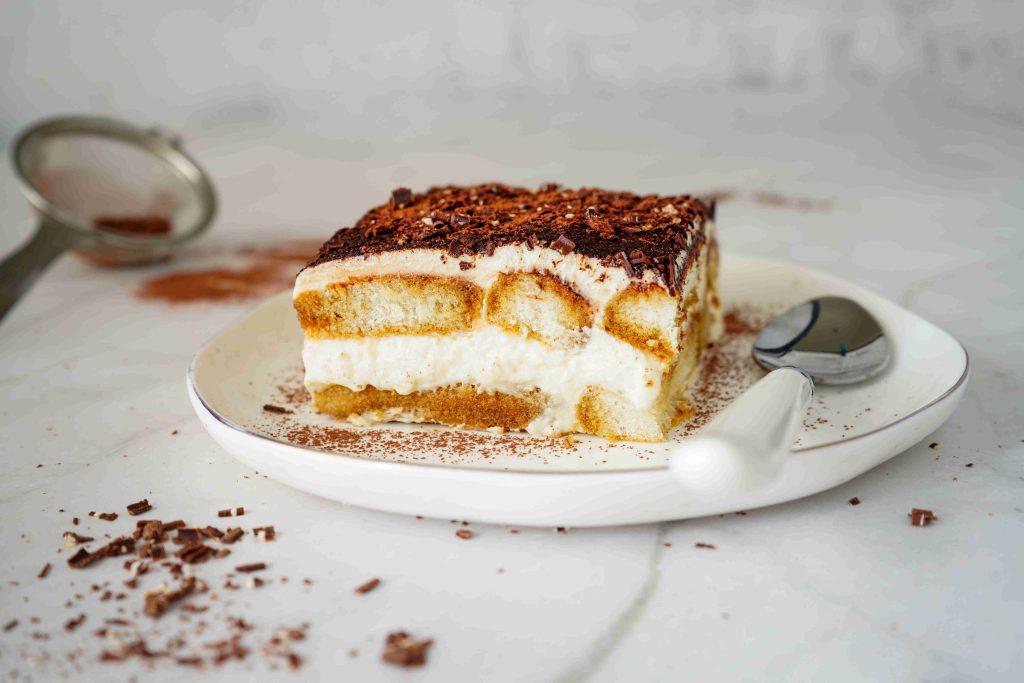 A slice of Christmas tiramisu topped with chocolate shavings sits on a white plate with a spoon, on a marble surface. A sifter and scattered chocolate pieces are in the background.
