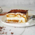 A slice of Christmas tiramisu topped with chocolate shavings sits on a white plate with a spoon, on a marble surface. A sifter and scattered chocolate pieces are in the background.