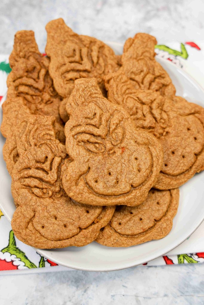 A plate of Grinch themed cookies, each shaped like the Grinch’s face and decorated with detailed features, sits on a festive napkin adorned in holiday colors.