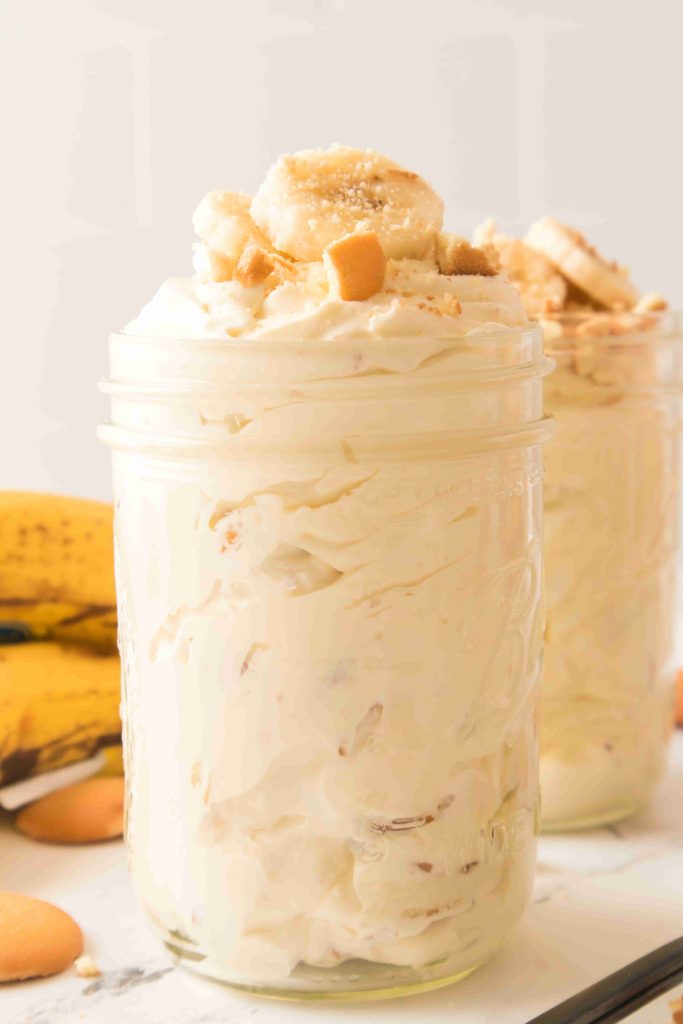 A mason jar filled with creamy Christmas Banana Pudding, topped with banana slices and vanilla wafer crumbs, sits on a white surface with bananas and vanilla wafers in the background.