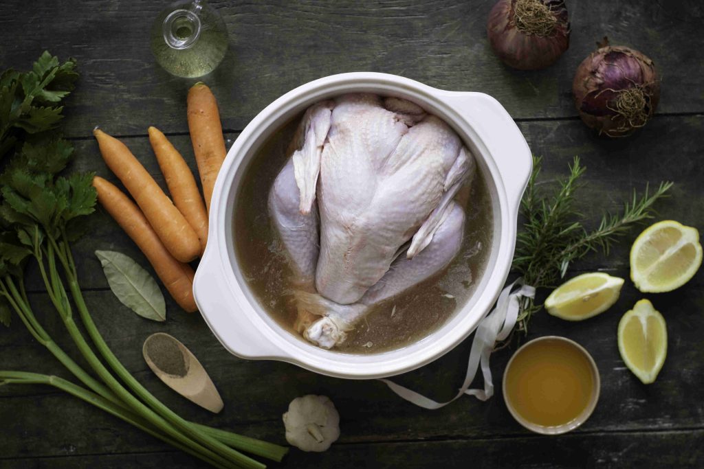 A raw whole chicken sits in a pot of broth, surrounded by carrots, celery, lemon halves, onions, fresh herbs, garlic, pepper, bay leaves—perfect inspiration for your next Holiday Turkey feast—alongside cooking oil and a bowl of broth on a dark wooden surface.
