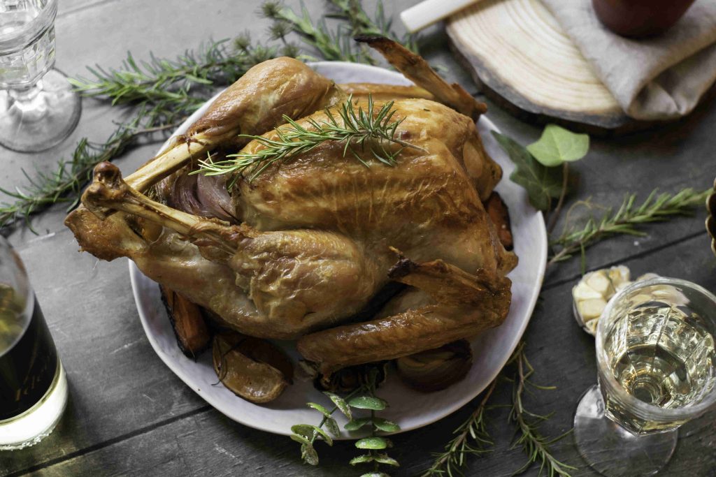 A roasted whole chicken garnished with fresh rosemary sits on a white plate, surrounded by herbs and glasses of white wine on a rustic wooden table, creating a festive scene reminiscent of a Holiday Turkey feast.