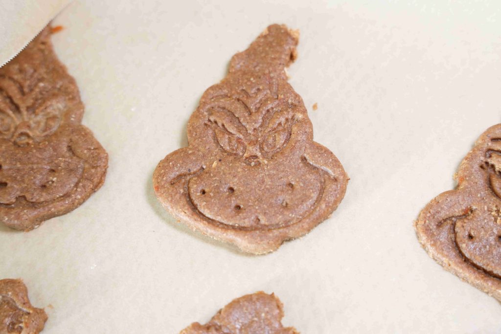 Grinch themed cookies made of chocolate cookie dough, shaped like the Grinch with his signature smirk and facial features, rest on parchment paper ready for baking.