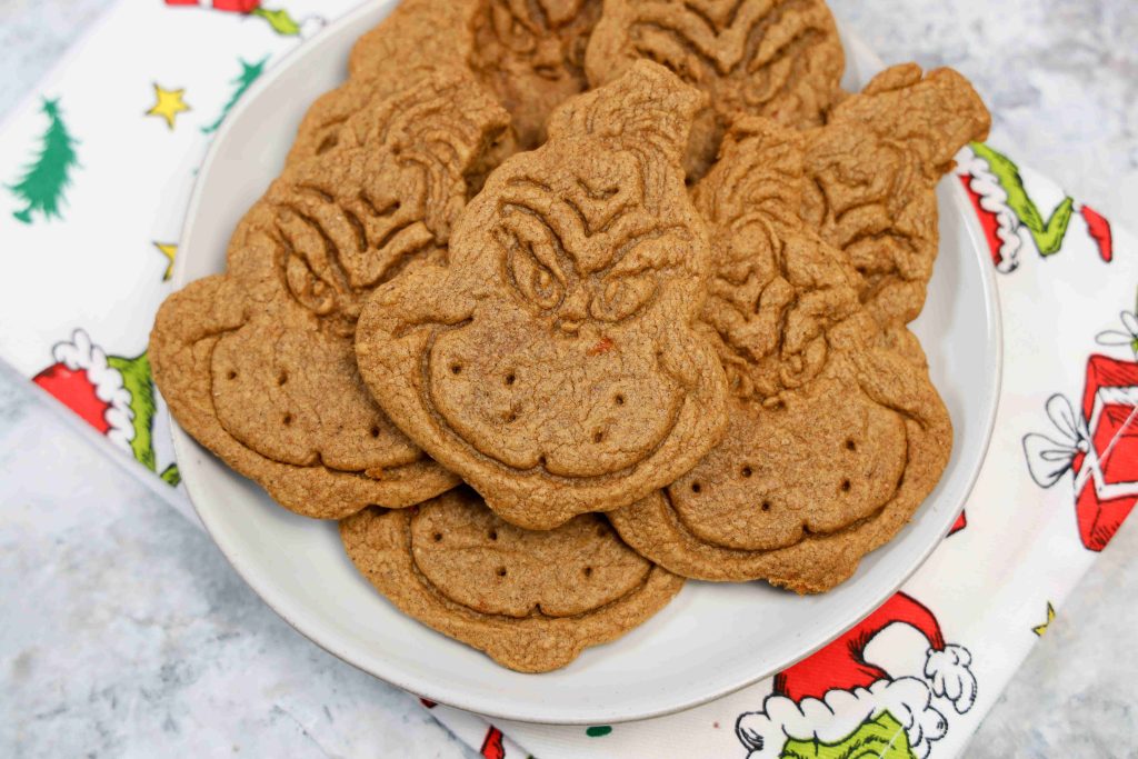 A plate of Grinch themed cookies shaped and decorated like the Grinch, placed on a festive holiday napkin adorned with Christmas tree and present designs.
