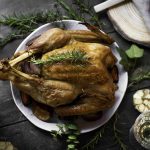 A roasted whole chicken garnished with rosemary sits on a white plate, surrounded by herbs, garlic, glassware, and napkins—creating a charming scene reminiscent of a holiday turkey feast on a rustic dark wooden table.