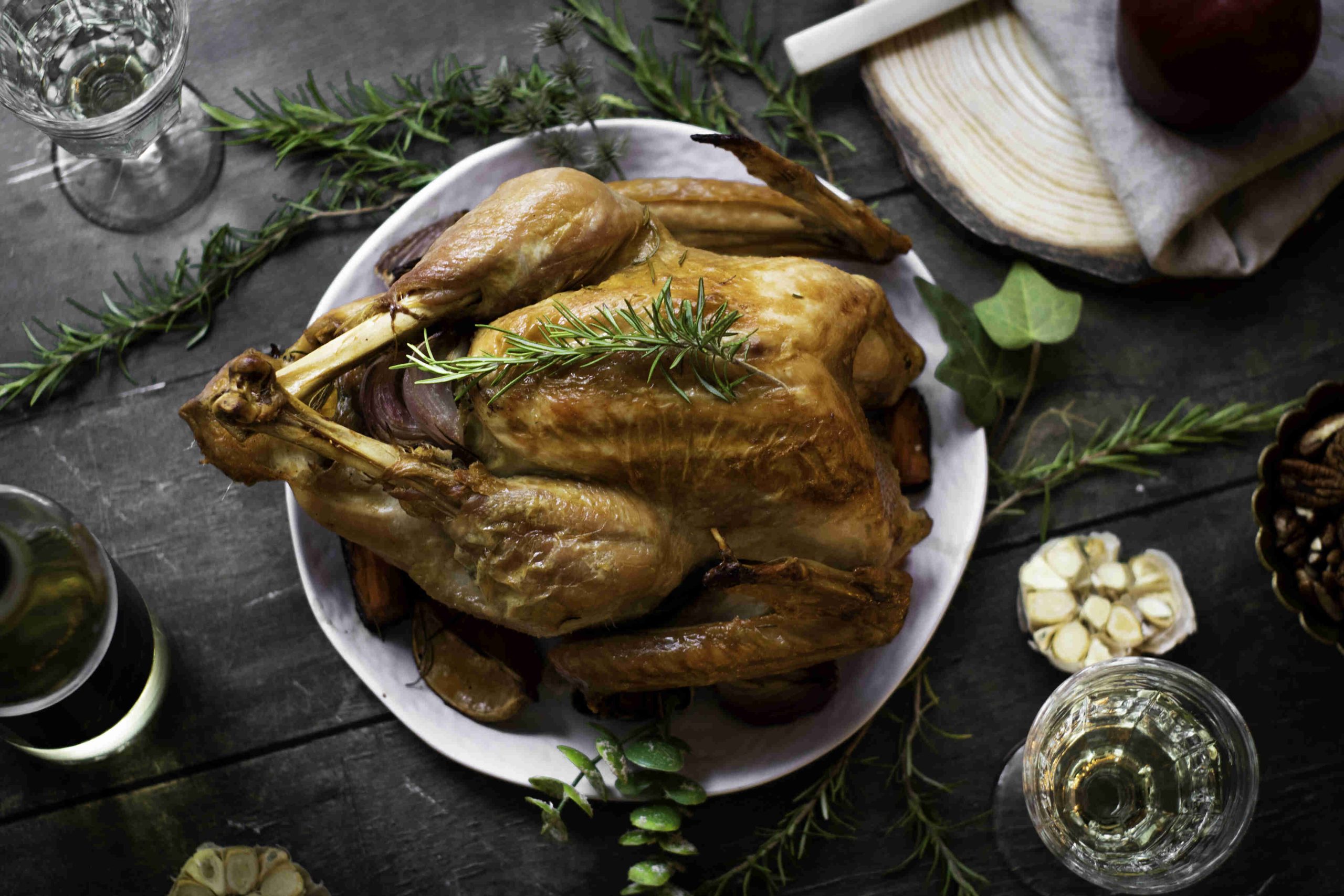 A roasted whole chicken garnished with rosemary sits on a white plate, surrounded by herbs, garlic, glassware, and napkins—creating a charming scene reminiscent of a holiday turkey feast on a rustic dark wooden table.