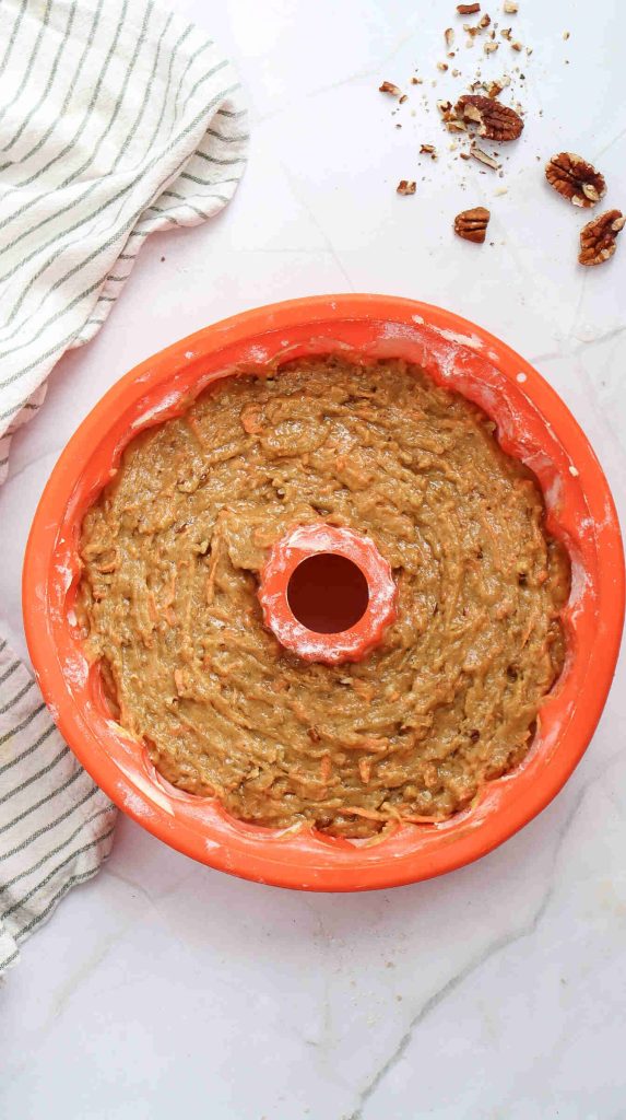 A silicone Bundt pan filled with unbaked Christmas Carrot Cake batter sits on a marble surface, next to a striped cloth and a few scattered pecans.