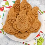 A plate of grinch themed cookies shaped like the Grinch’s face sits on a napkin decorated with the Grinch, Christmas trees, and presents. The cookies are golden brown with detailed facial features.