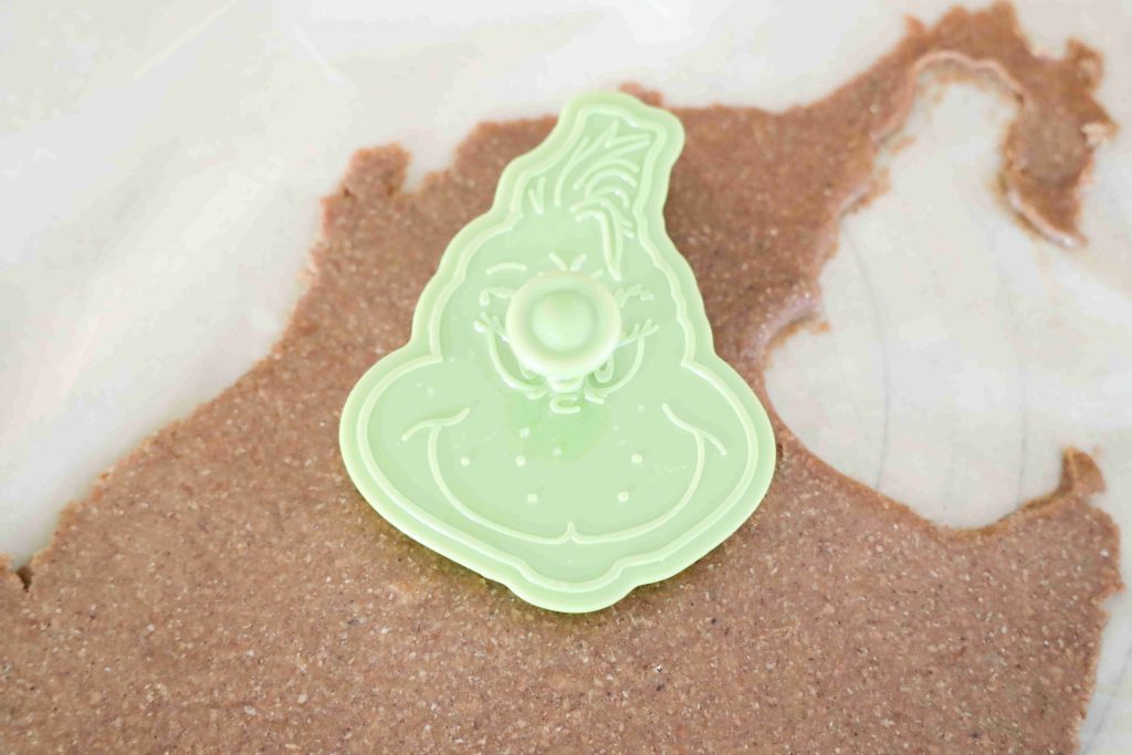 A green cookie cutter shaped like a cartoon character is placed on rolled-out brown dough, perfect for making Grinch themed cookies on a light surface.