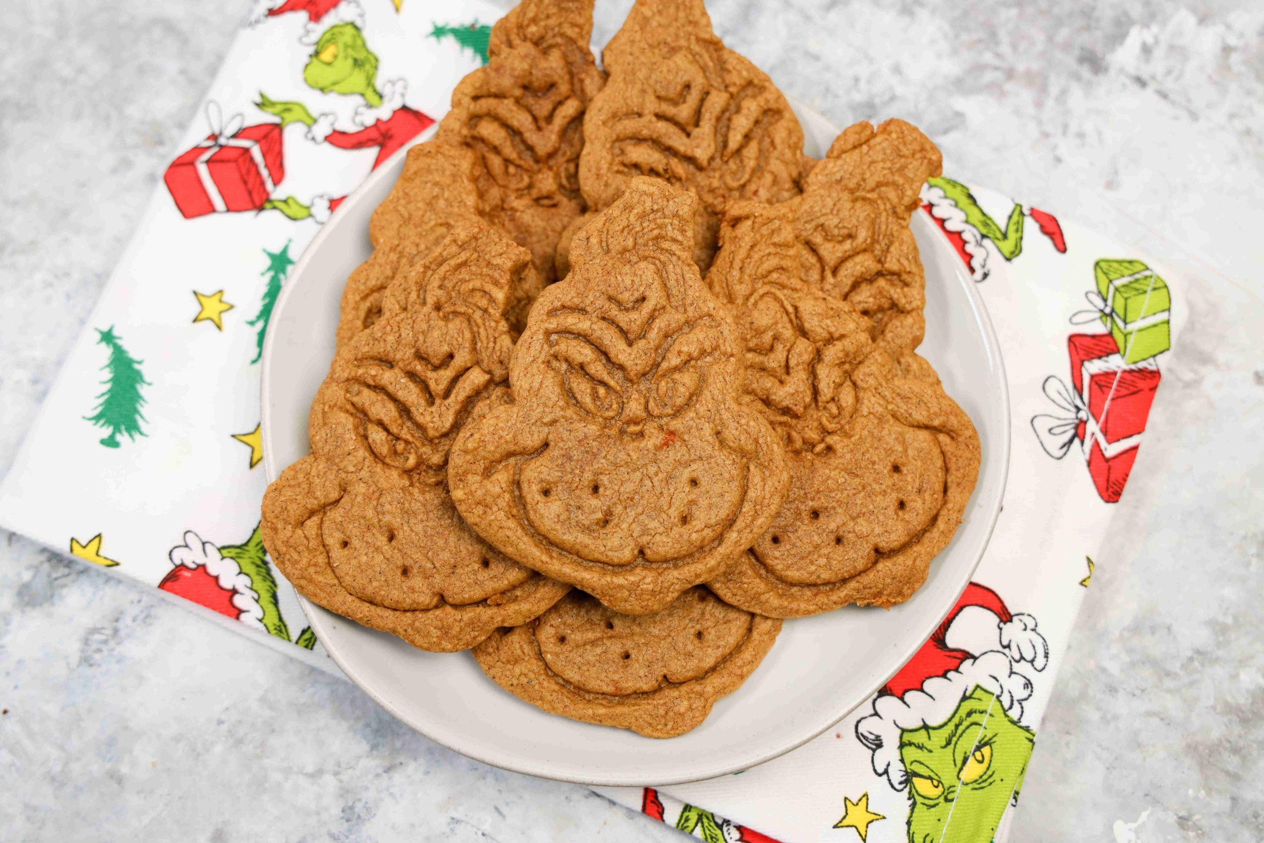 A plate of grinch themed cookies shaped like the Grinch’s face sits on a napkin decorated with the Grinch, Christmas trees, and presents. The cookies are golden brown with detailed facial features.