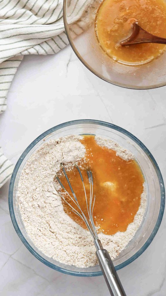 Two glass bowls on a white surface: one with flour, a whisk, and liquid being mixed for Christmas Carrot Cake, and the other with a wet mixture and a wooden spoon. A striped dish towel is partially visible in the corner.