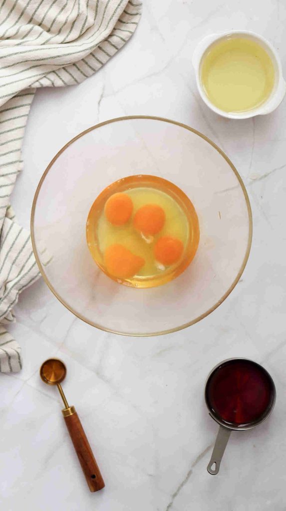 A glass bowl with three whole eggs and one egg yolk, ready for Christmas Carrot Cake, surrounded by a small bowl of oil, a striped towel, a metal measuring cup of liquid, and a tablespoon on a marble countertop.