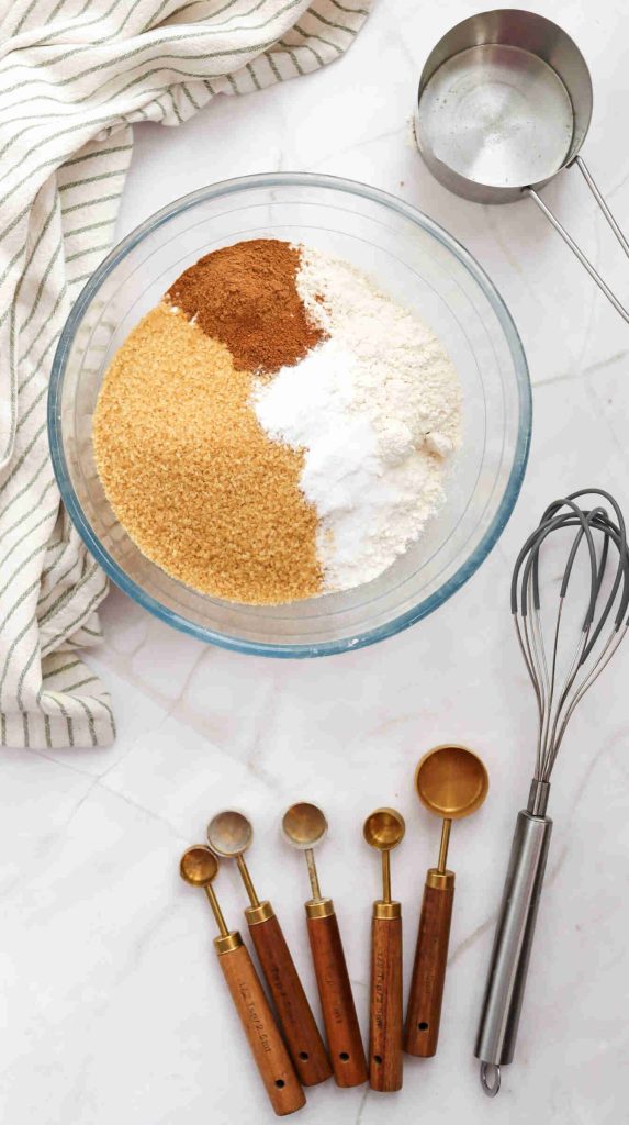 A glass bowl containing flour, brown sugar, cinnamon, and baking powder sits on a marble surface next to a whisk, measuring spoons, and a striped kitchen towel—perfect for preparing Christmas Carrot Cake.