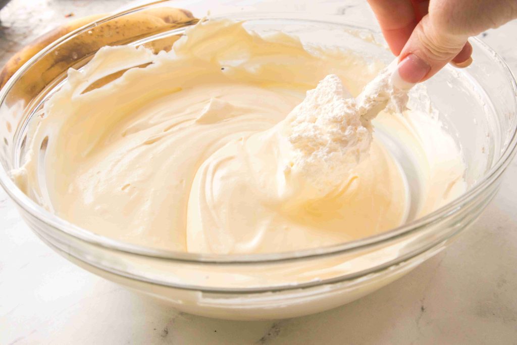 A hand holding a spatula mixes creamy, whipped Christmas Banana Pudding batter in a clear glass bowl on a light-colored countertop.