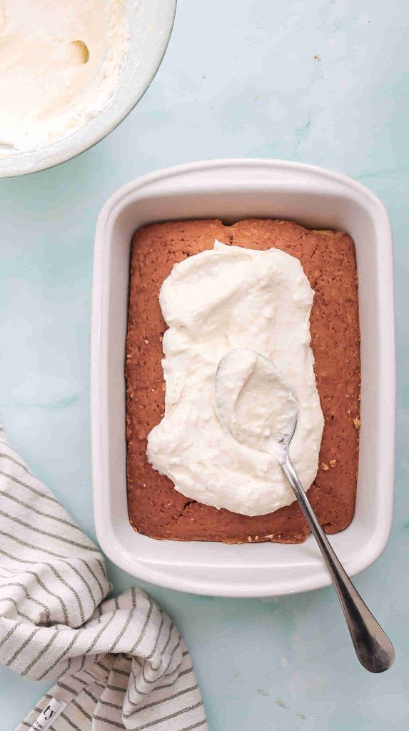 A coconut Christmas cake in a white dish is being topped with whipped cream using a spoon. A mixing bowl with leftover cream and a striped cloth are nearby on a light blue surface.