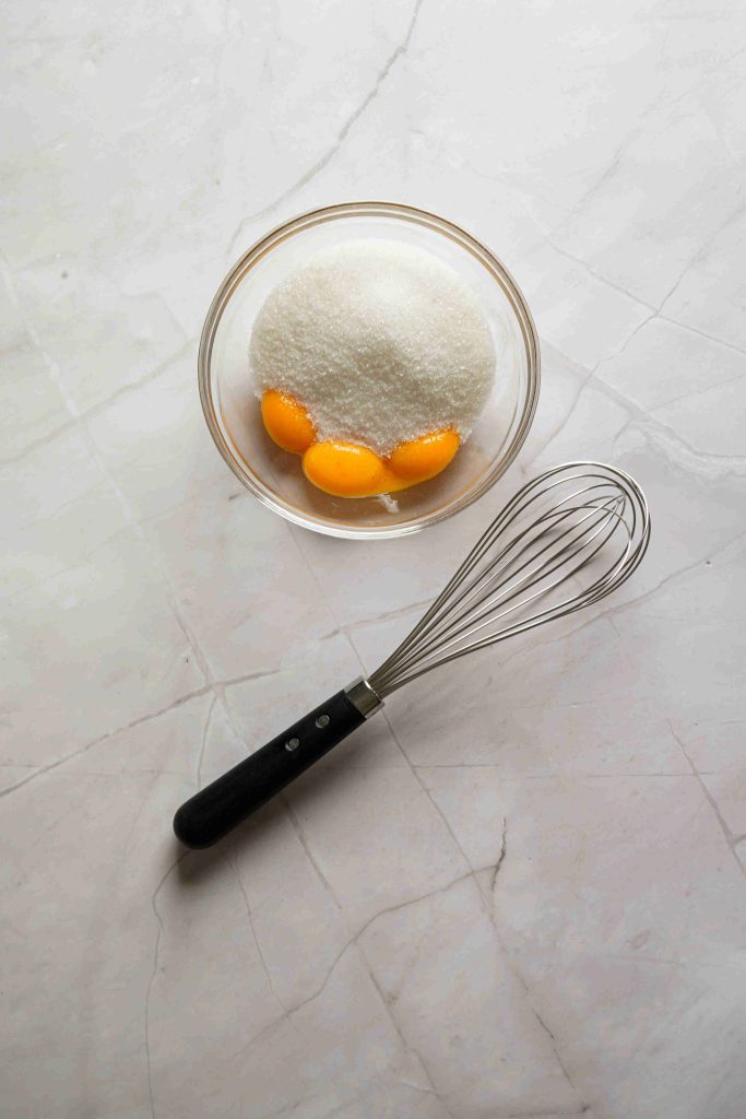 A glass bowl with three egg yolks and granulated sugar on a light marble surface, next to a metal whisk with a black handle—perfect for starting your Christmas tiramisu.