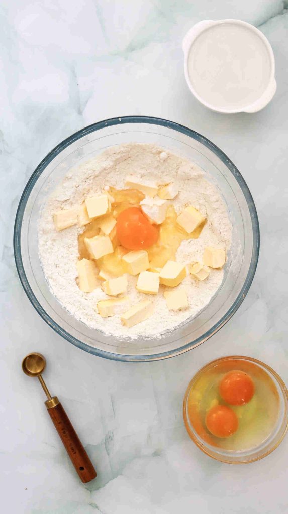 A glass bowl with flour, cubed butter, and an egg in the center for coconut Christmas cake; nearby are a bowl with two eggs, a measuring spoon, and a small bowl of milk on a marble surface.
