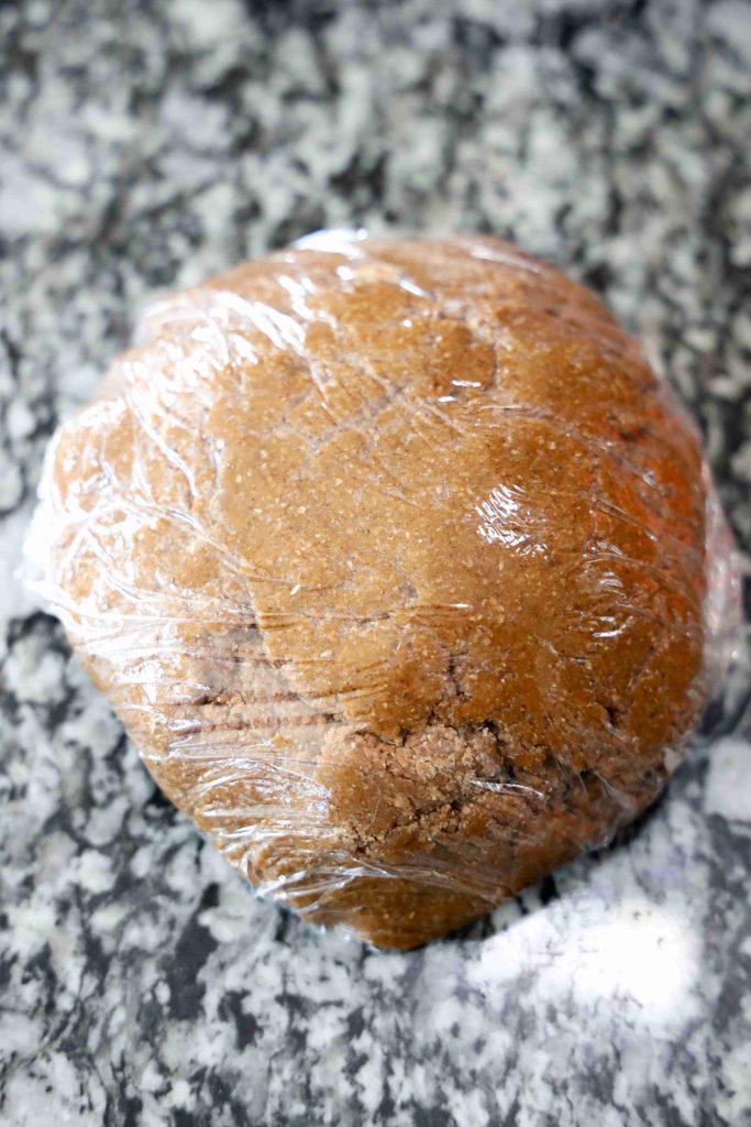 A ball of brown dough for grinch themed cookies is wrapped in plastic wrap and sits on a marbled countertop.