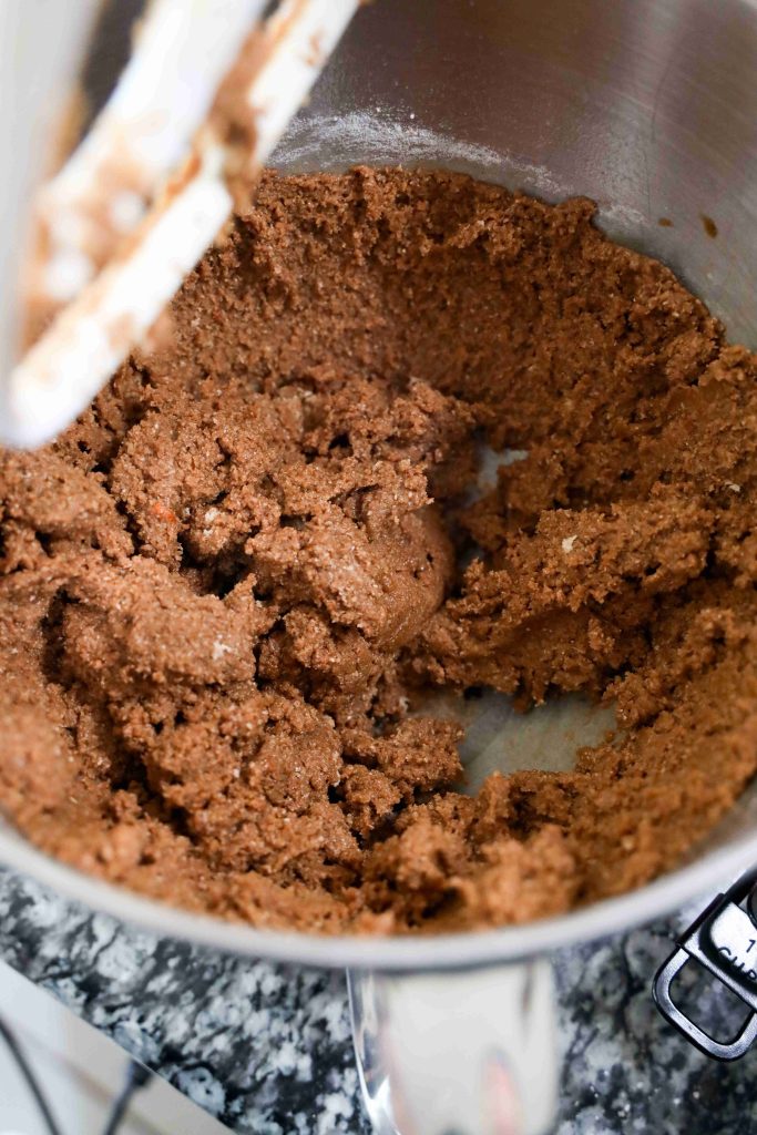 Chocolate cookie dough for grinch themed cookies is being mixed in a silver stand mixer bowl, with some dough clinging to the paddle attachment above. The mixer sits on a granite countertop.