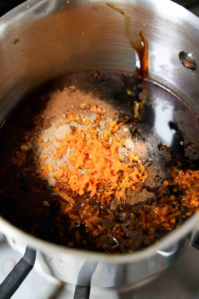 A close-up of a stainless steel pot on a stove containing dark liquid, orange zest, and assorted spices, ready to be mixed or cooked for grinch themed cookies.
