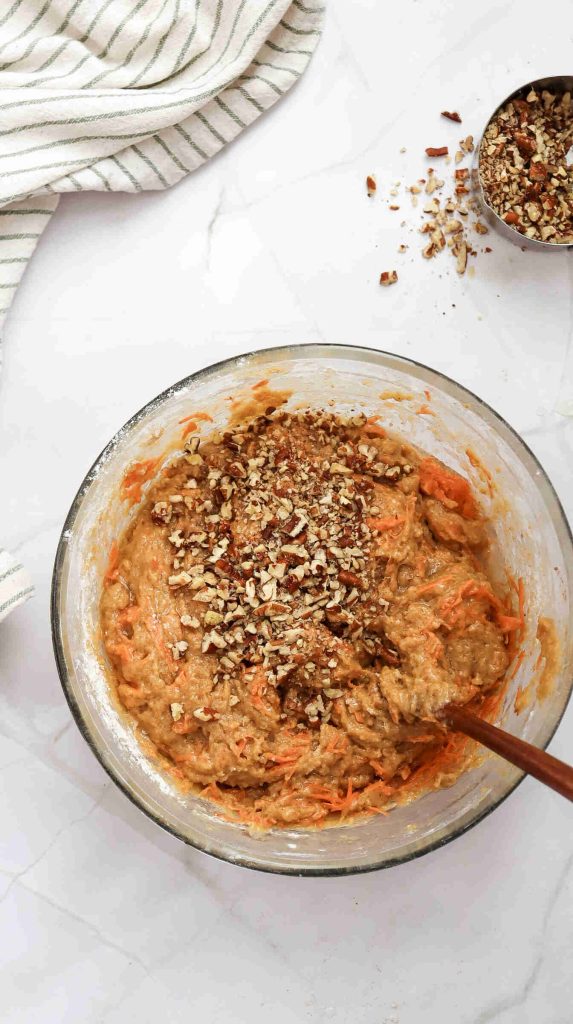 A glass bowl filled with Christmas Carrot Cake batter topped with chopped nuts, being mixed with a wooden spoon. A striped cloth and a measuring cup with more chopped nuts are nearby on a white countertop.