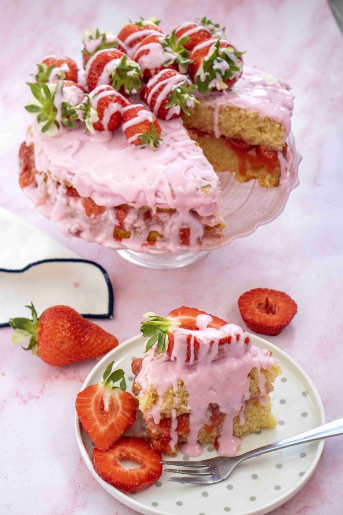 A valentine's day cake with pink icing and whole strawberries sits on a cake stand. A slice has been cut and served on a plate with fresh strawberries, all against a light pink background.