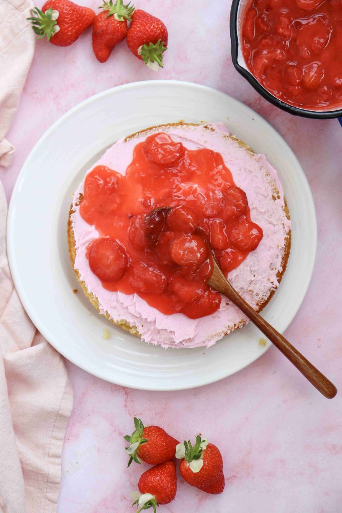 Overhead view of a Valentine's Day cake topped with pink frosting and strawberry sauce, with a wooden spoon spreading the sauce. Fresh strawberries and a bowl of extra sauce sit nearby on a pink surface.