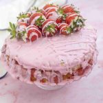 A pink frosted Valentine's Day cake topped with fresh strawberries and drizzled with white icing sits on a cake stand. Strawberry pieces show through its creamy, textured layers, with a white cloth in the background.